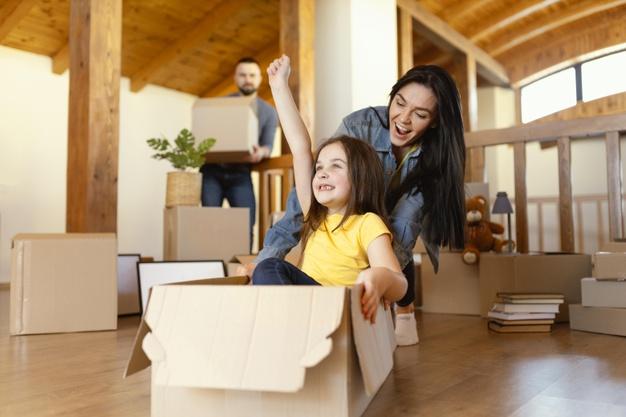 Familia feliz por la mudanza a su nuevo hogar soñado mientras el marido de fondo se encuentra cargando cajas