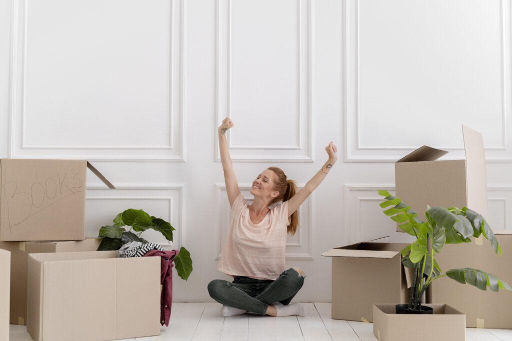 Chica disfrutando de su casa soñada mientras al lado tiene cajas de mudanzas para hacer el traslado a su nueva casa
